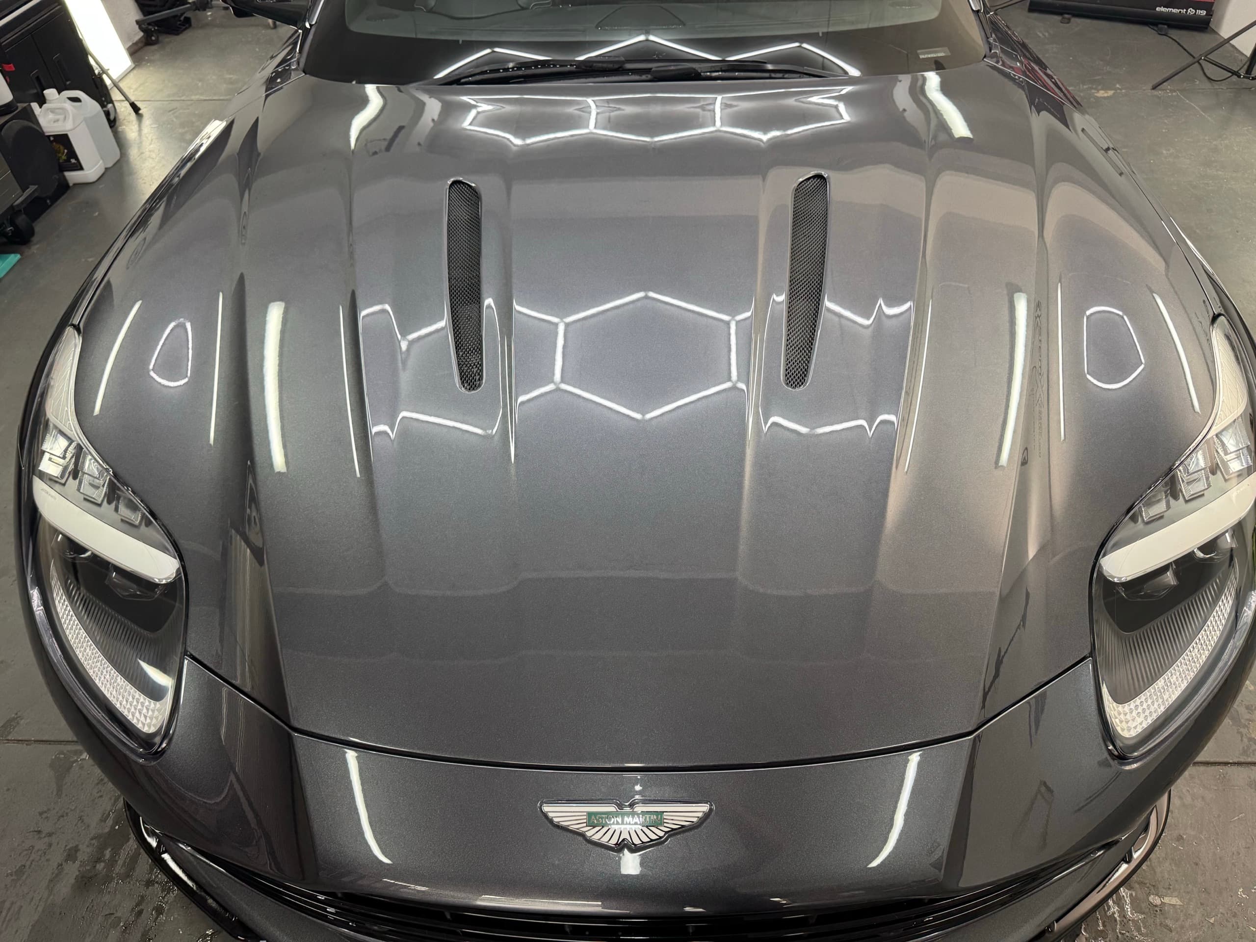 Front of a grey Aston Martin DB12 with hexagonal studio lights reflected across the bonnet