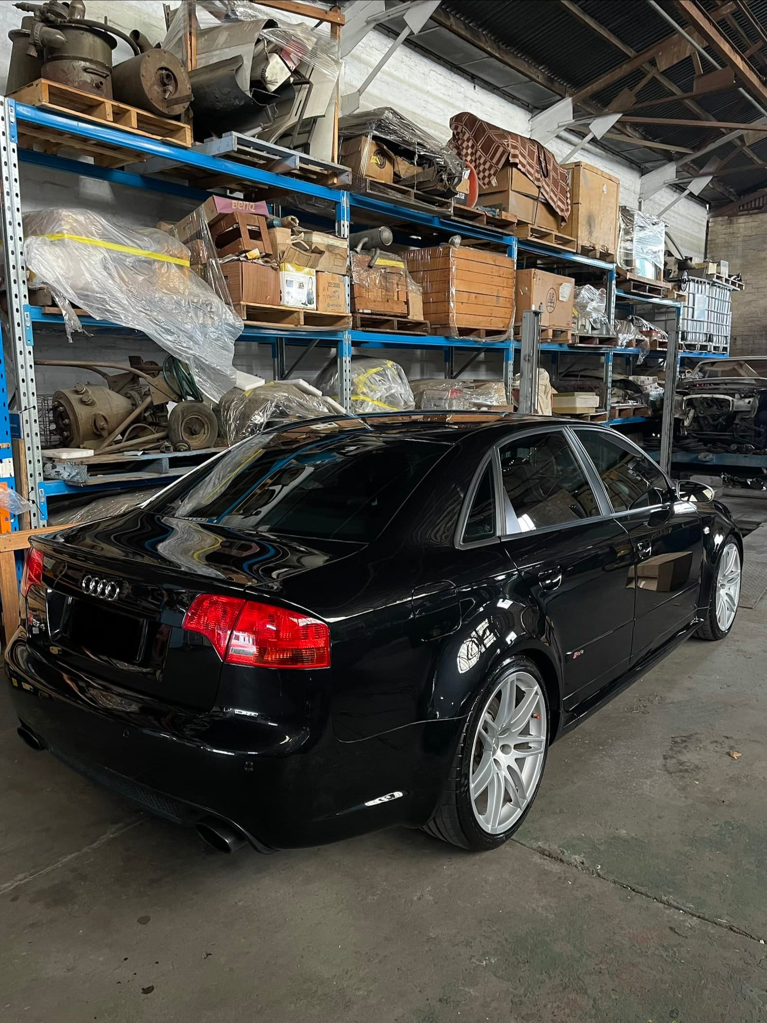 Rear three-quarter view of a black Audi RS4 sedan in a dimly lit warehouse, with light reflected along its glossy paint