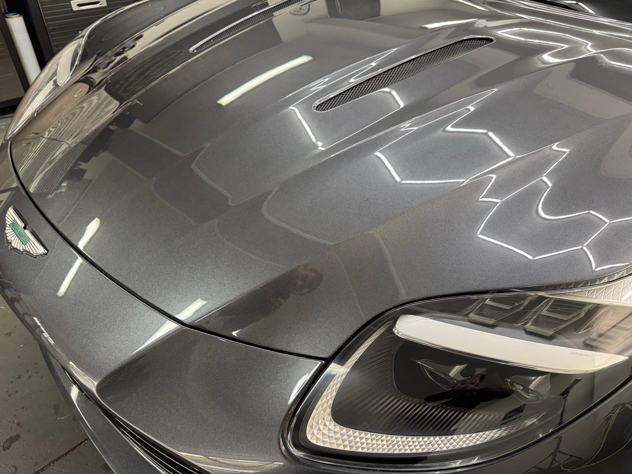 Close-up of a grey Aston Martin bonnet, badge and headlight with studio lights reflected in the paint