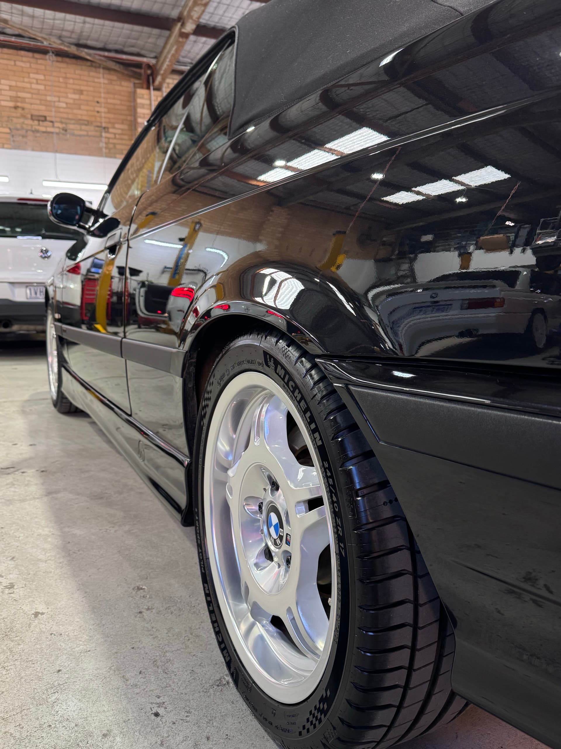 Low-angle view along a black BMW E36 M3 with a silver alloy wheel and workshop lights reflected in the paint