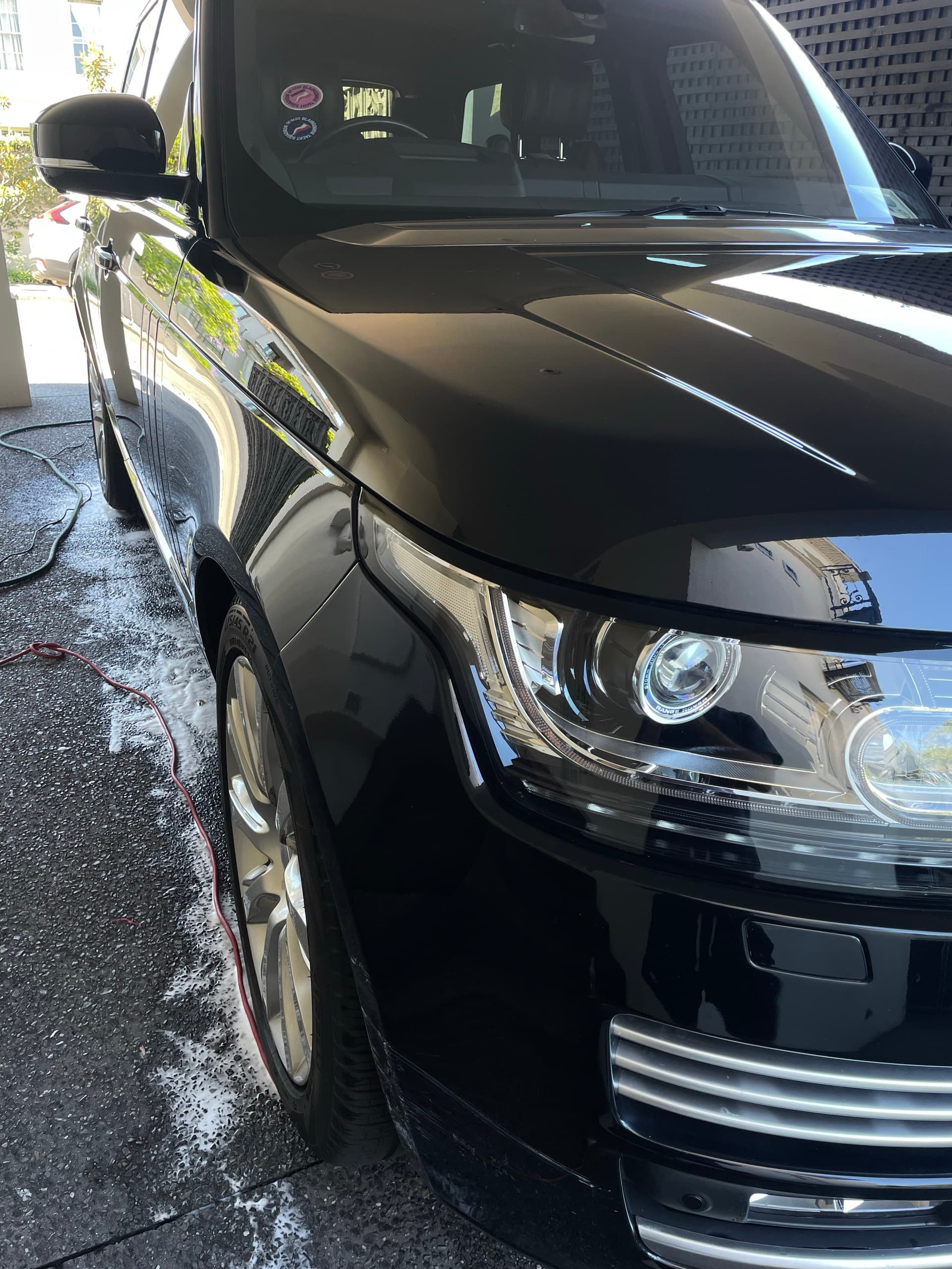 Front corner and headlight of a black Range Rover, wet from washing, with foam on the driveway